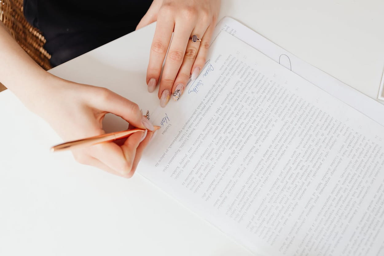 Een hand schrijft met een gouden pen op een stapel papieren met tekst. De vrouw heeft verzorgde nagels en draagt een ring.