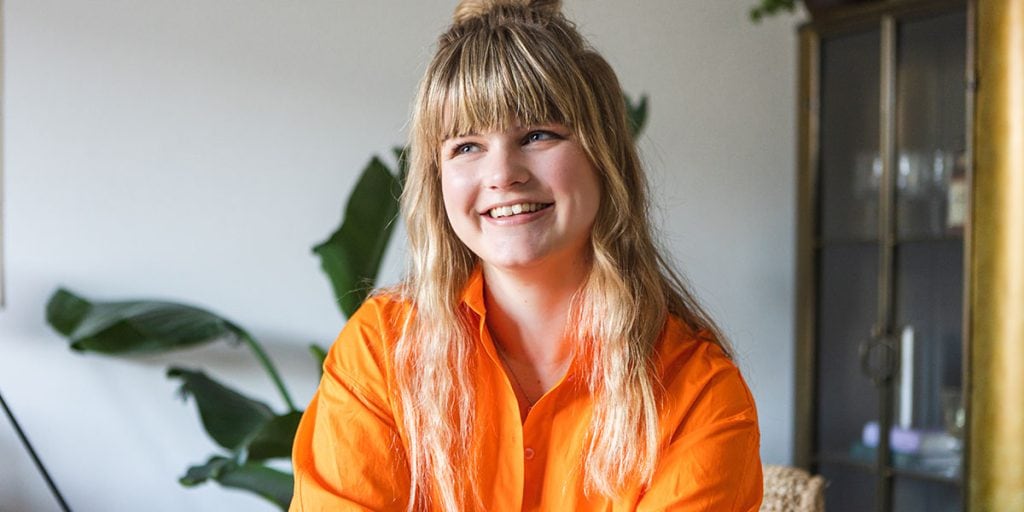 Een lachende vrouw met lang, golvend haar draagt een feloranje blouse en zit in een lichte, moderne ruimte met groene planten op de achtergrond.