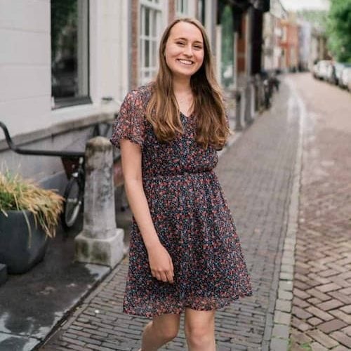 Jonge vrouw in een bloemenjurk wandelt glimlachend over een straat met historische gebouwen en fietsen. De sfeer is vrolijk en uitnodigend, perfect voor mode- en lifestyle-inhoud.