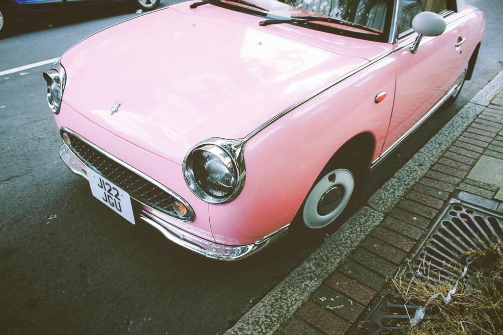 Roze retro auto geparkeerd aan de straat met een klassieke uitstraling en chromen details. Het voertuig heeft een opvallende kleur en een schattig ontwerp dat de aandacht trekt.