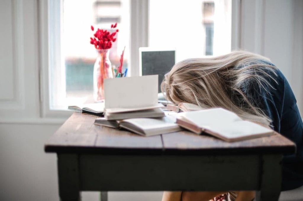 Een vrouw met lang haar leunt moeizaam op een bureau vol open boeken, terwijl een vaas met rode bloemen op de achtergrond staat.