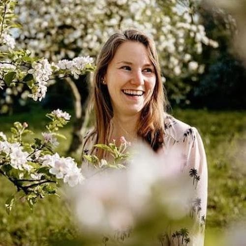 Vrouw die lacht te midden van bloeiende bomen in een zonnige tuin. De frisse bloemen en groene achtergrond creëren een vrolijke en natuurlijke sfeer.