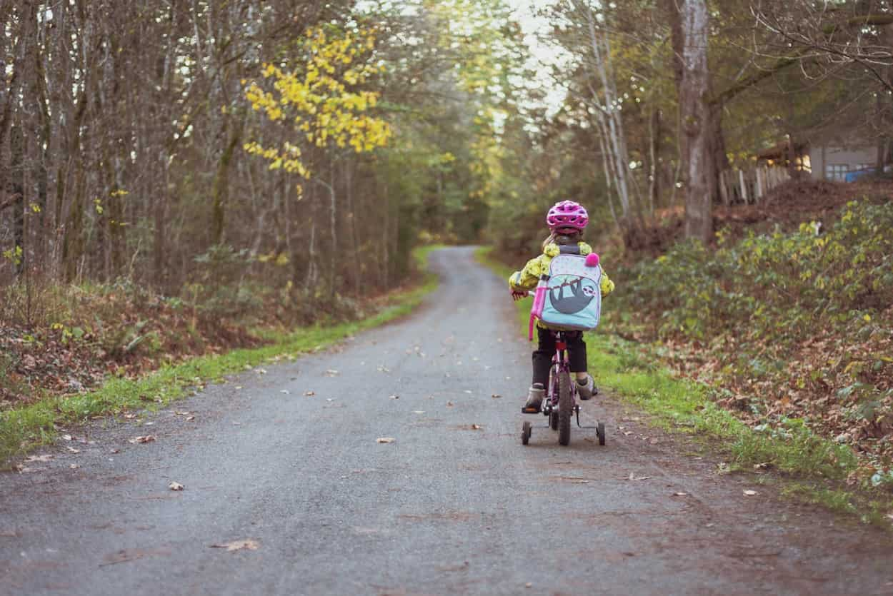 Een meisje fietst op een smal pad omringd door bomen, met een kleurrijke rugzak en een roze fietshelm.