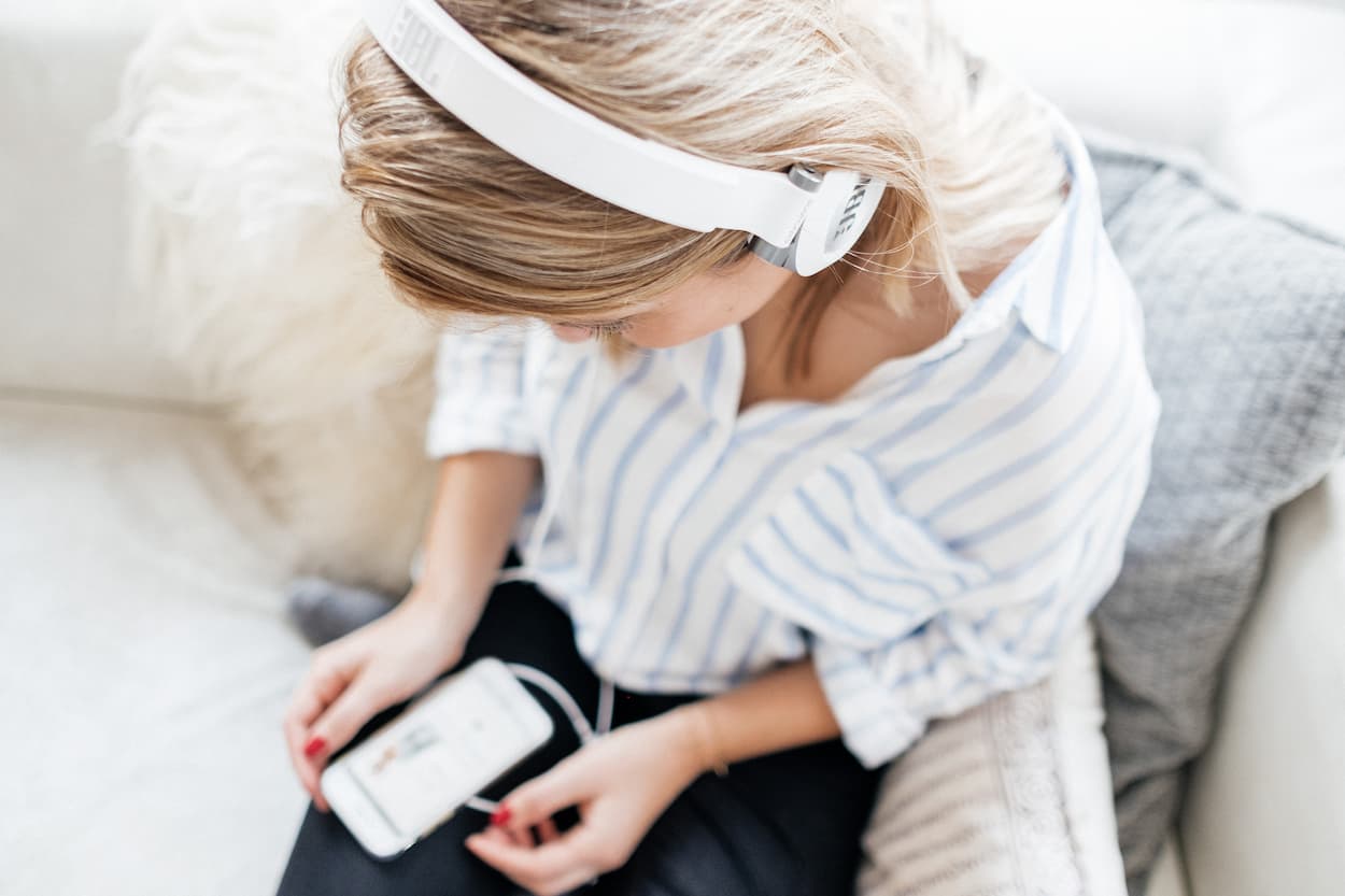 Een vrouw met een witte koptelefoon luistert naar muziek op haar smartphone terwijl ze op een bank zit.