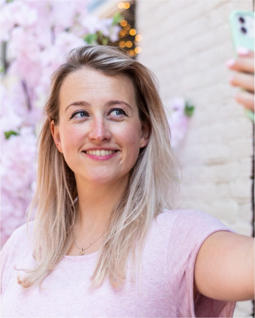 Jonge vrouw met lang, lichtblond haar maakt een selfie in een sfeervolle omgeving met bloesem en zachte verlichting. Haar glimlach straalt vreugde en zelfvertrouwen uit.