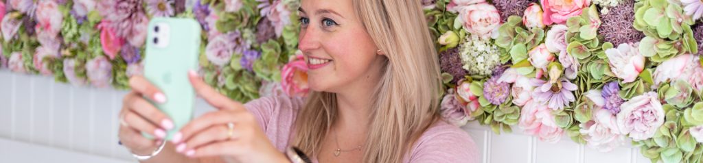 Vrouw met lang, blond haar maakt een selfie voor een kleurrijke bloemenachtergrond. De afbeelding straalt een vrolijke en positieve sfeer uit.