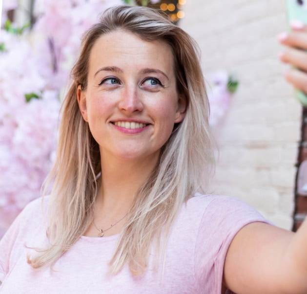 Jonge vrouw maakt een selfie met een smartphone, omringd door bloesembomen. Ze lacht en kijkt blij in de camera.