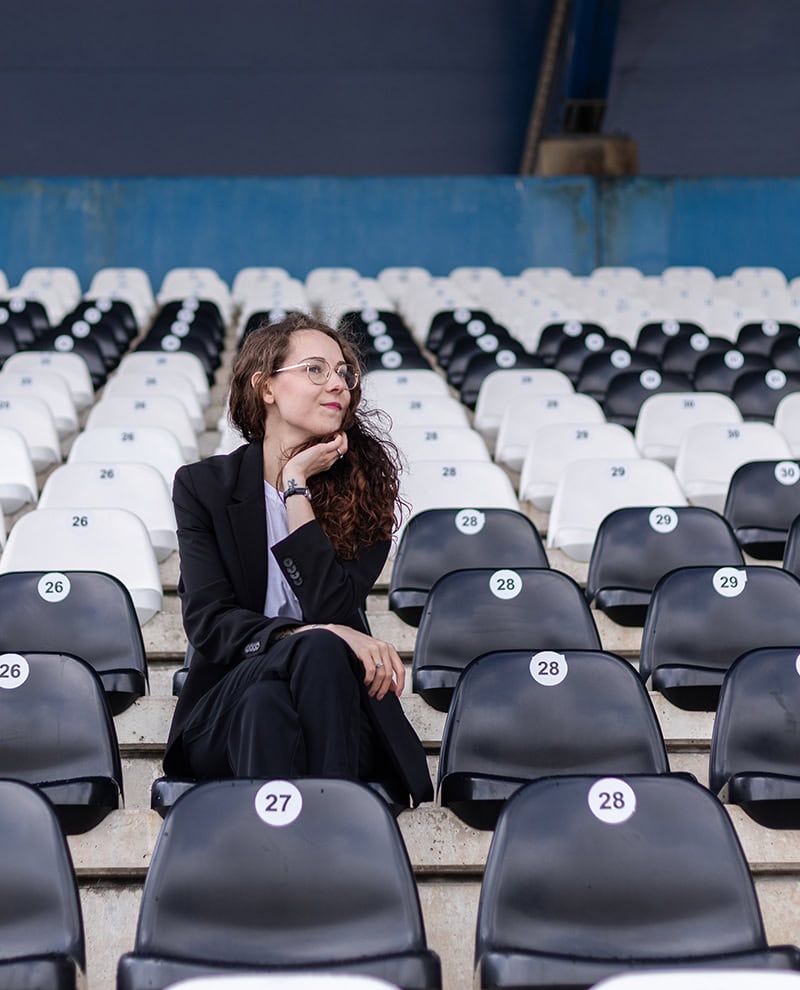 Een vrouw in een zwart pak zit pensief op de tribune van een stadion, omringd door lege zwart-witte stoelen. Haar uitstraling straalt zelfvertrouwen en reflectie uit.