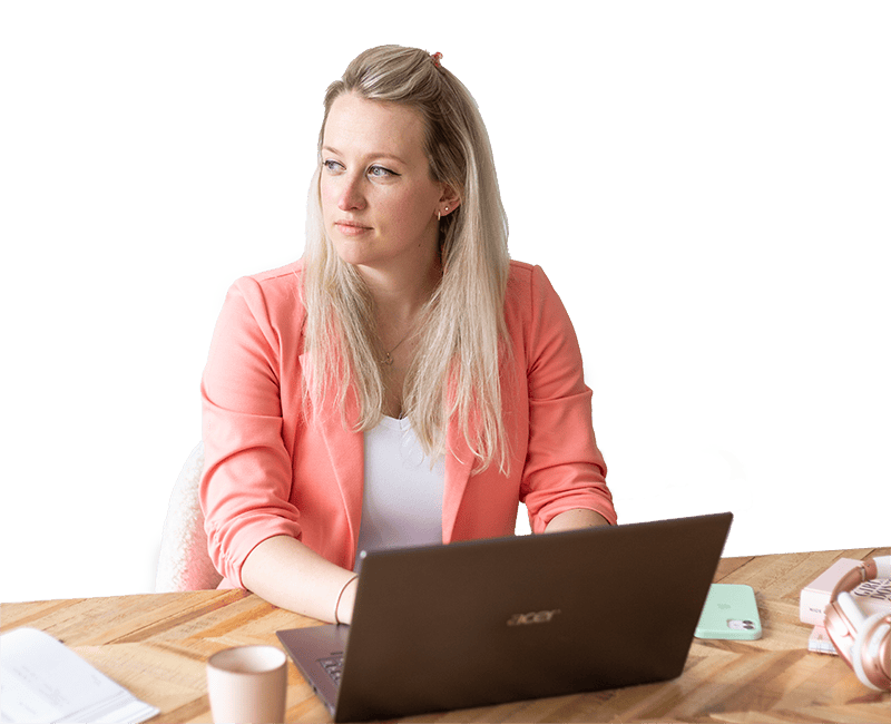 Een vrouw met lang, blond haar zit achter een laptop op een houten tafel. Ze draagt een roze blazer en kijkt geconcentreerd in de richting van het scherm, met een kopje koffie en andere kantoorbenodigdheden op de tafel.