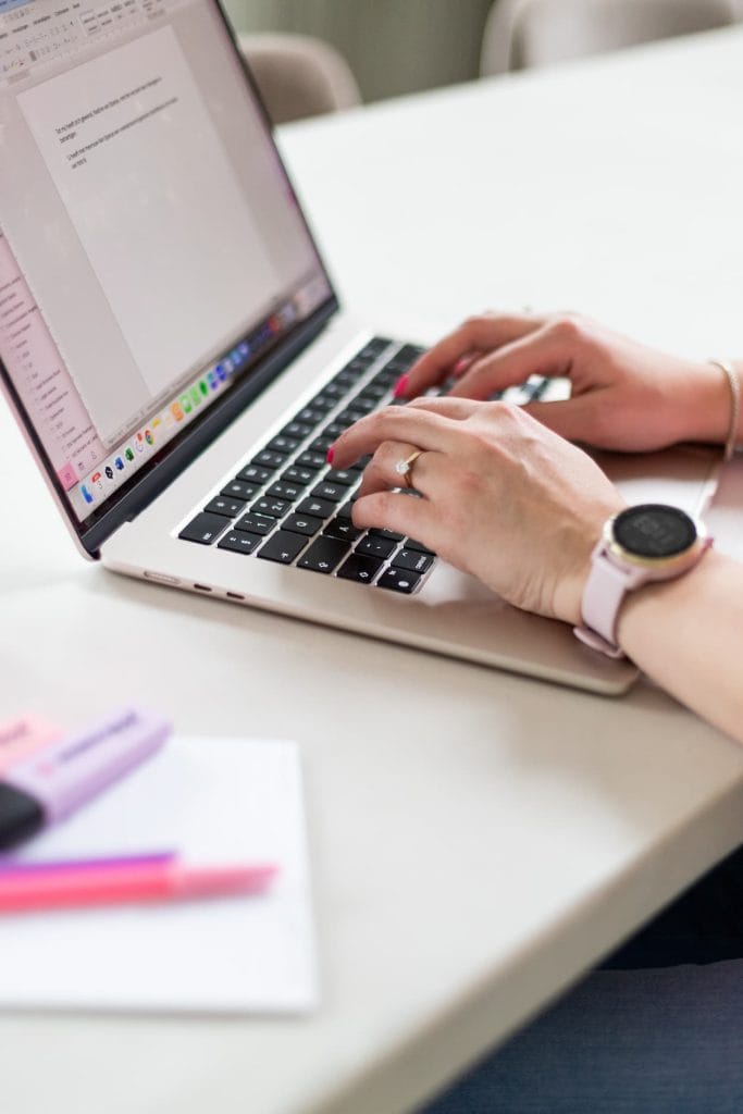 Een vrouw typt op een laptop met een witte achtergrond, omringd door pennen en notitiepapier. De focus ligt op haar handen en de laptop, wat een productieve werkomgeving benadrukt.