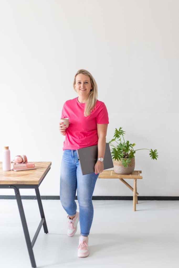 Een vrouw in een roze T-shirt houdt een koffiekopje vast en draagt een laptop onder haar arm, terwijl ze in een lichte, moderne ruimte staat. Op de tafel liggen een waterfles, koptelefoon en notitieboekje.