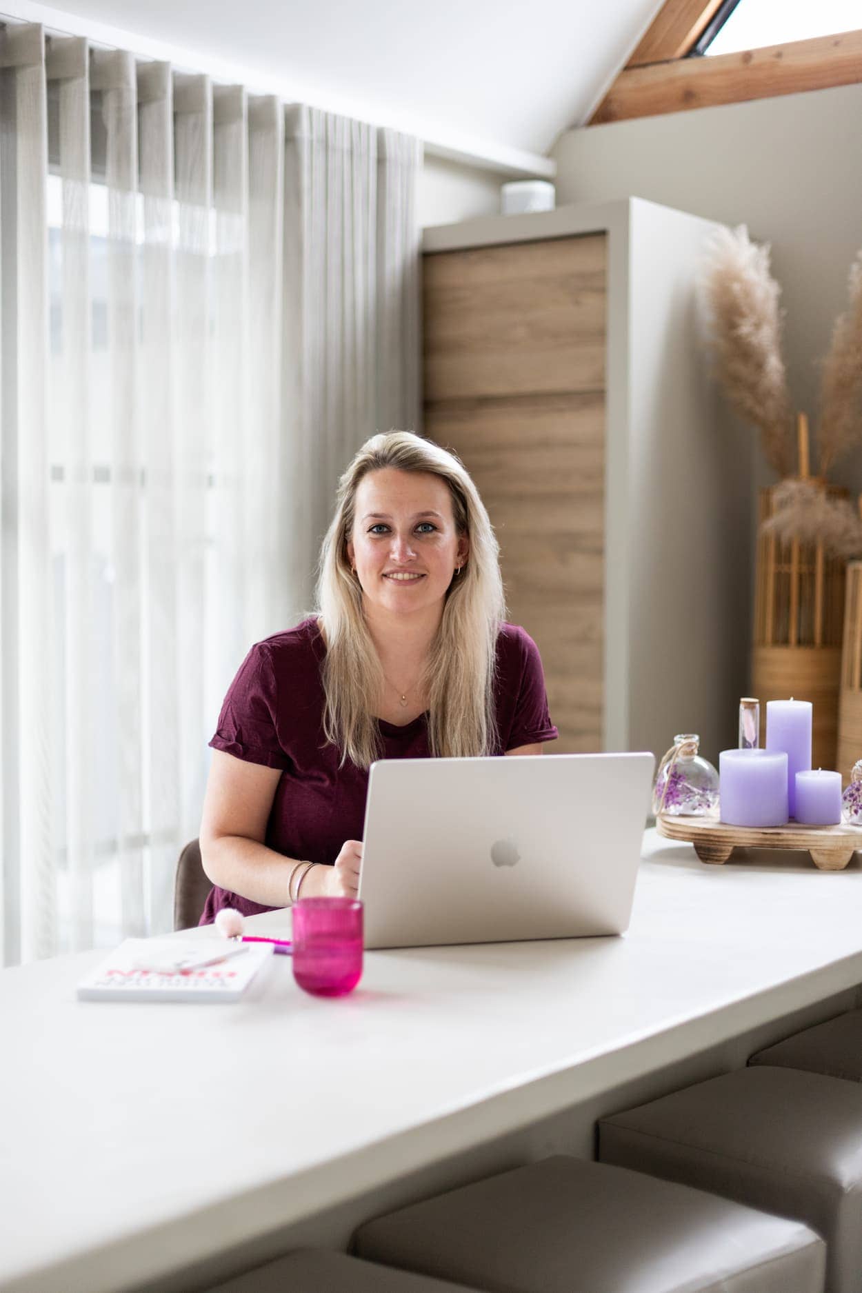 Een vrouw met lang blond haar zit aan een bureau met een laptop, omringd door een gezellige en lichte interieur. Op de tafel staan paarse kaarsen en een roze glas, wat een warme sfeer creëert.