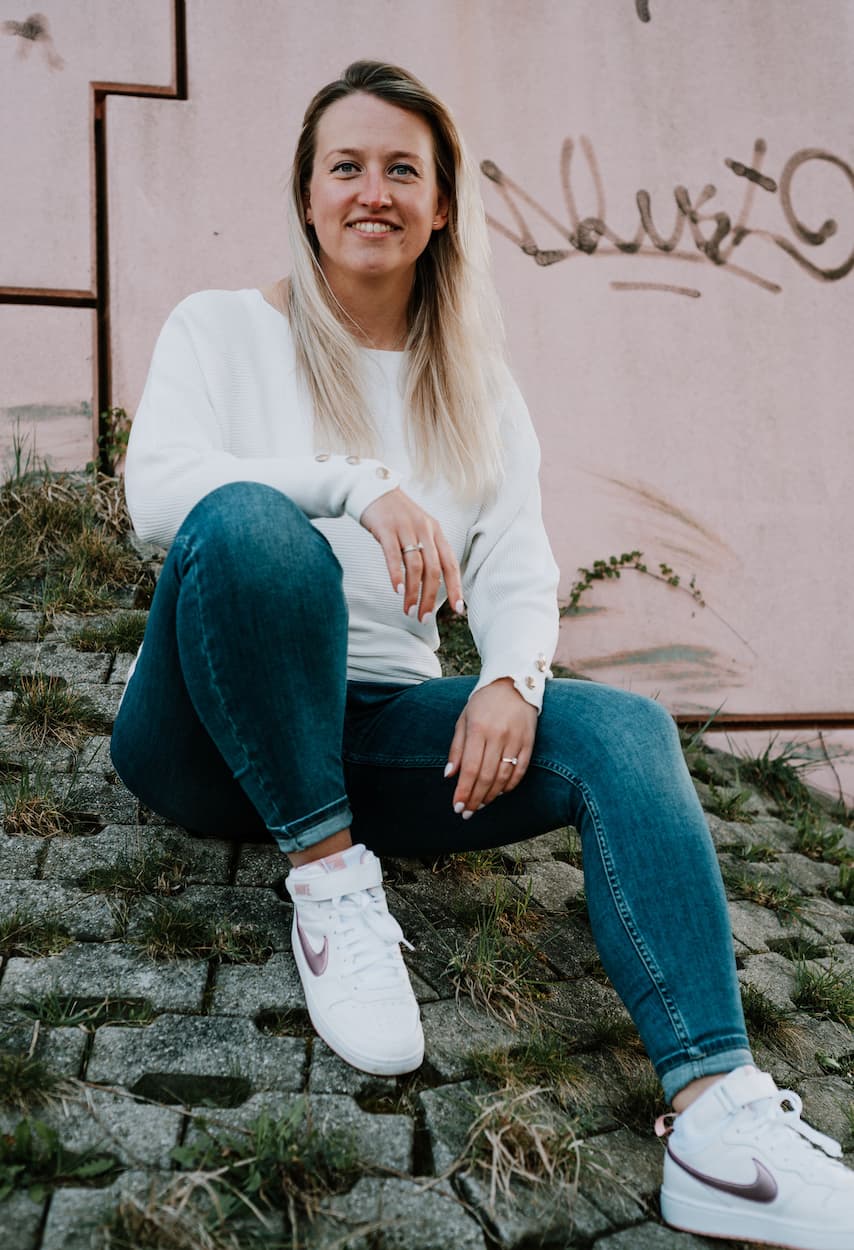 Vrouw in een casual outfit met een witte trui en spijkerbroek, zittend op een stenen ondergrond met gras. De achtergrond bevat een roze muur met graffiti, wat een stedelijke sfeer creëert.