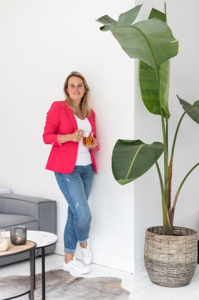 Vrouw in een roze blazer staat ontspannen met een glas thee in de hand, leunend tegen een witte muur naast een grote groene plant. De moderne woonkamer heeft een stijlvolle uitstraling met een grijze bank en een ronde salontafel.