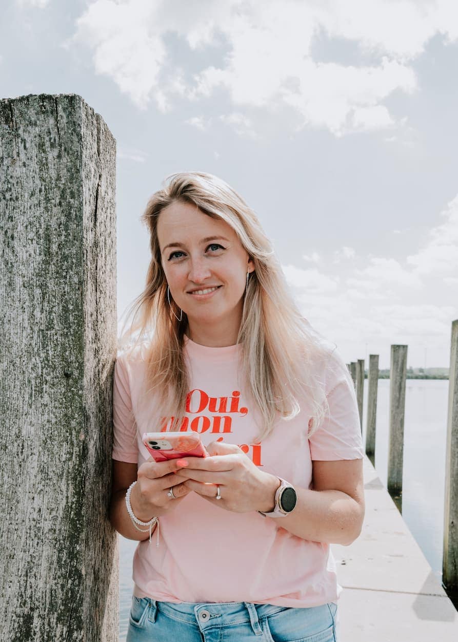 Jonge vrouw met lang blond haar draagt een lichtroze T-shirt en kijkt glimlachend naar haar telefoon terwijl ze leunt tegen een houten paal aan de waterkant.