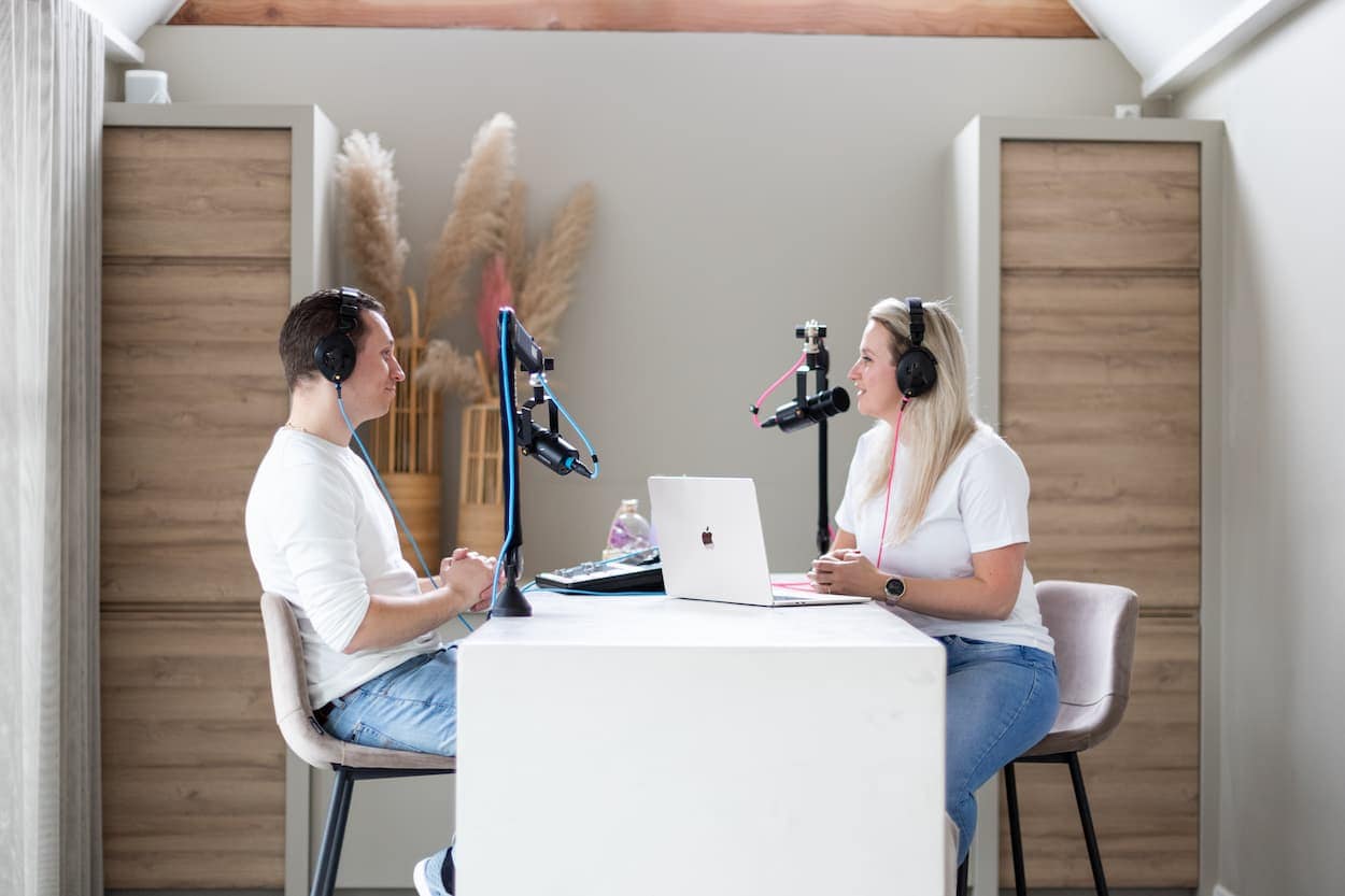 Twee mensen in een podcaststudio, met microfoons en een laptop op een witte tafel. De vrouw spreekt terwijl de man aandachtig luistert, omringd door een moderne en gezellige inrichting.