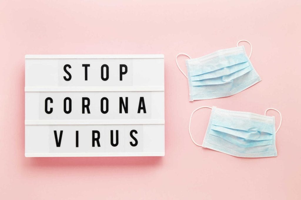 Wit bord met de tekst "STOP CORONA VIRUS" en twee blauwe mondmaskers op een roze achtergrond. Deze afbeelding benadrukt het belang van het dragen van mondmaskers tijdens de coronapandemie.