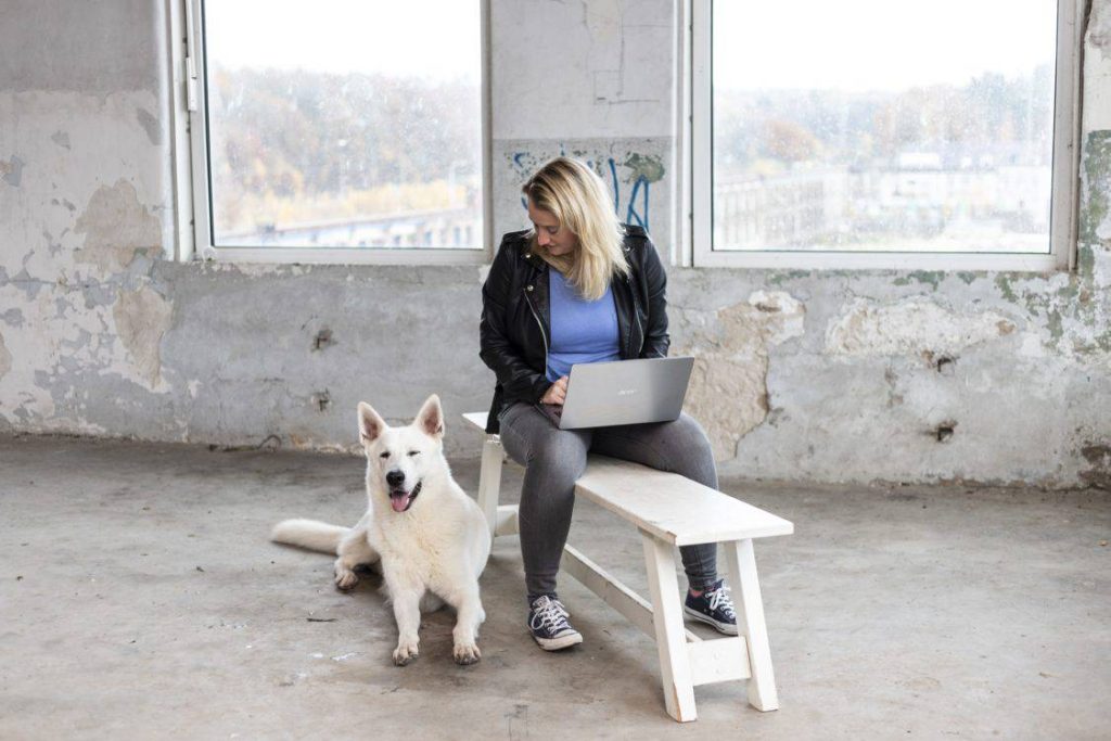 Een vrouw zit op een houten bankje in een verlaten ruimte met een laptop, terwijl een witte hond naast haar ligt. De ruimte heeft grote ramen met uitzicht op een buitenomgeving.