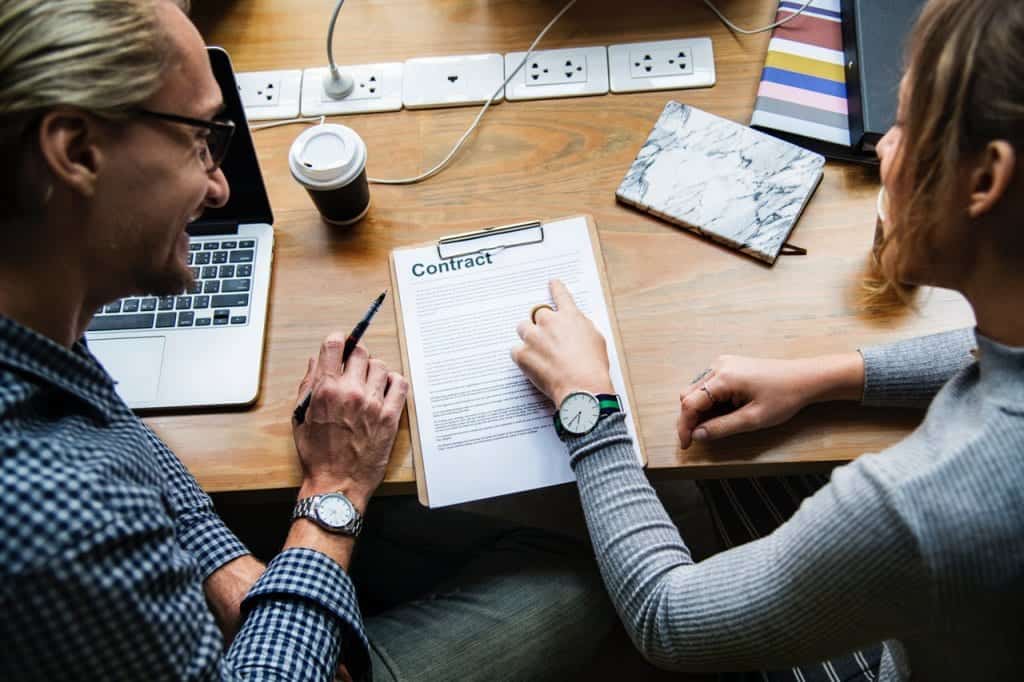 Twee mensen bespreken een contract aan een houten tafel, met een laptop en een koffiebeker op de achtergrond. De vrouw wijst naar het contract terwijl de man naar haar kijkt.