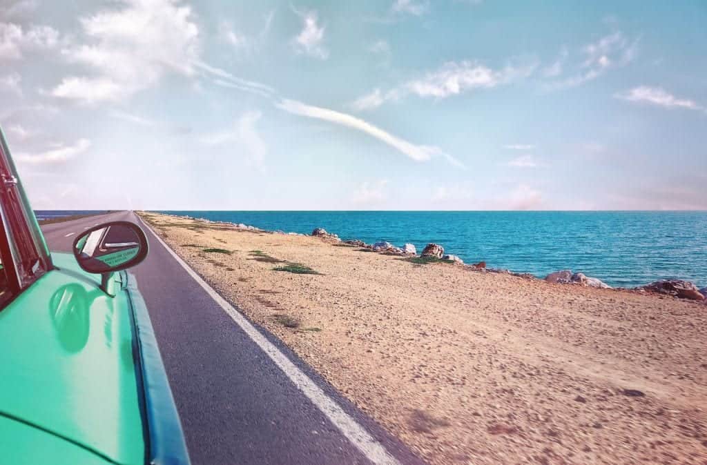 Een groene auto rijdt langs een kustweg met uitzicht op de oceaan en een heldere lucht. Het beeld straalt een gevoel van vrijheid en avontuur uit.