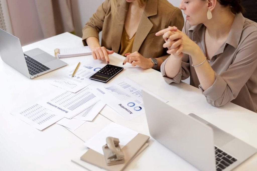 Twee vrouwen bespreken financiële gegevens en gebruiken een rekenmachine tijdens een zakelijke vergadering. Op de tafel liggen documenten en een laptop, wat de focus op samenwerking en analyse benadrukt.