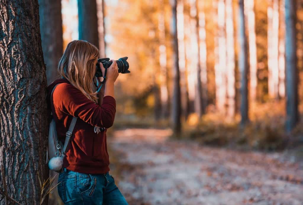 Een vrouw in een rode trui fotografeert in een bos met herfstkleuren, leunend tegen een boom. De achtergrond toont een prachtig herfstlandschap met vervaagde bomen.