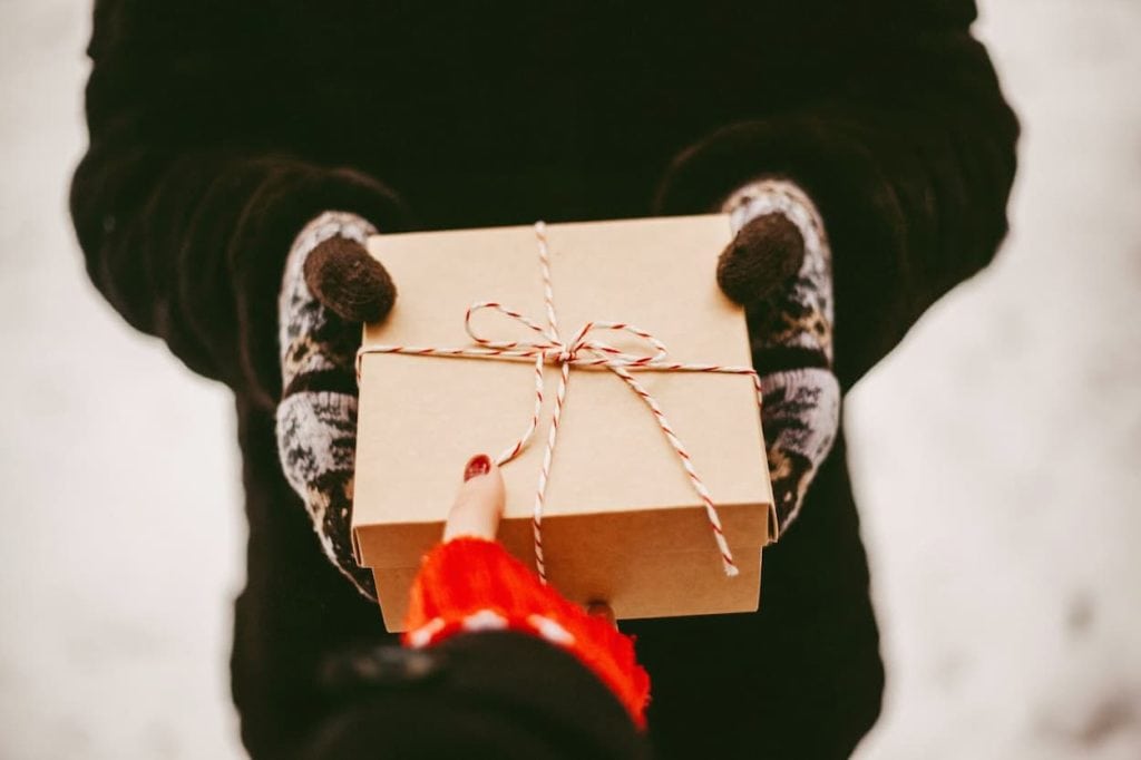 Een persoon in een zwarte jas overhandigt een mooi ingepakt cadeau met een rood-wit touwtje aan een andere persoon met een rode trui, terwijl de achtergrond bedekt is met sneeuw.