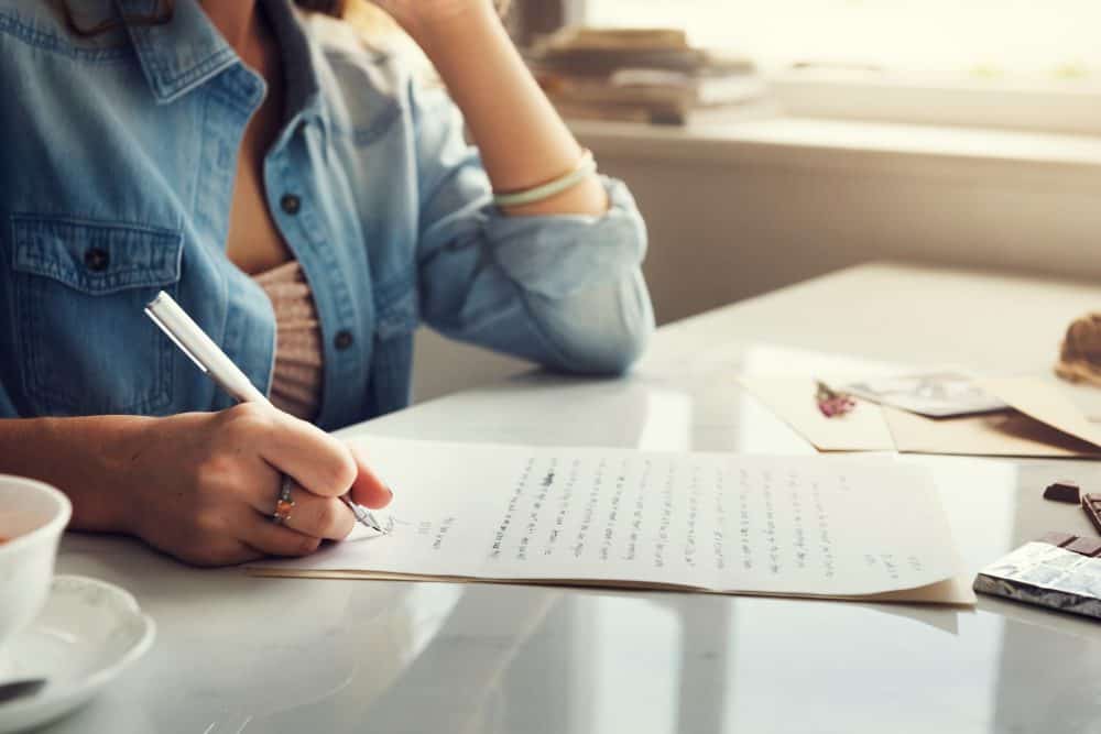 Een vrouw schrijft op een vel papier terwijl ze aan een tafel zit, omringd door foto's en een kopje koffie. De focus ligt op haar hand met een pen, wat de creativiteit en het schrijfproces benadrukt.