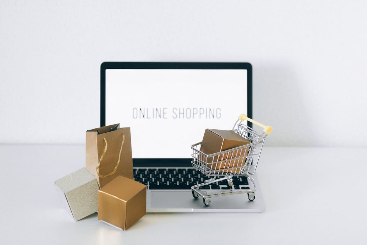 Laptop met het scherm dat "ONLINE SHOPPING" toont, omringd door een winkelwagentje en verschillende geschenkdozen. Dit beeld illustreert de moderne ervaring van online winkelen.