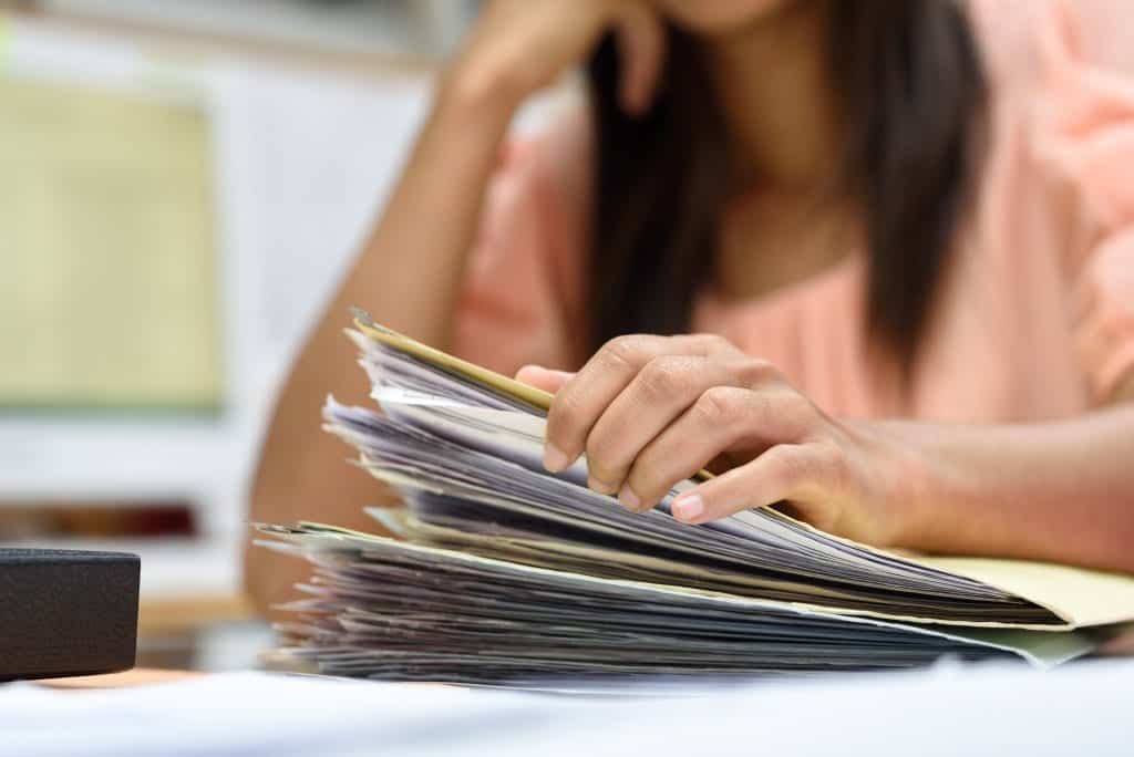 Een vrouwelijke hand die door een stapel documenten bladert op een bureau, wat een indruk geeft van organisatie en administratie.