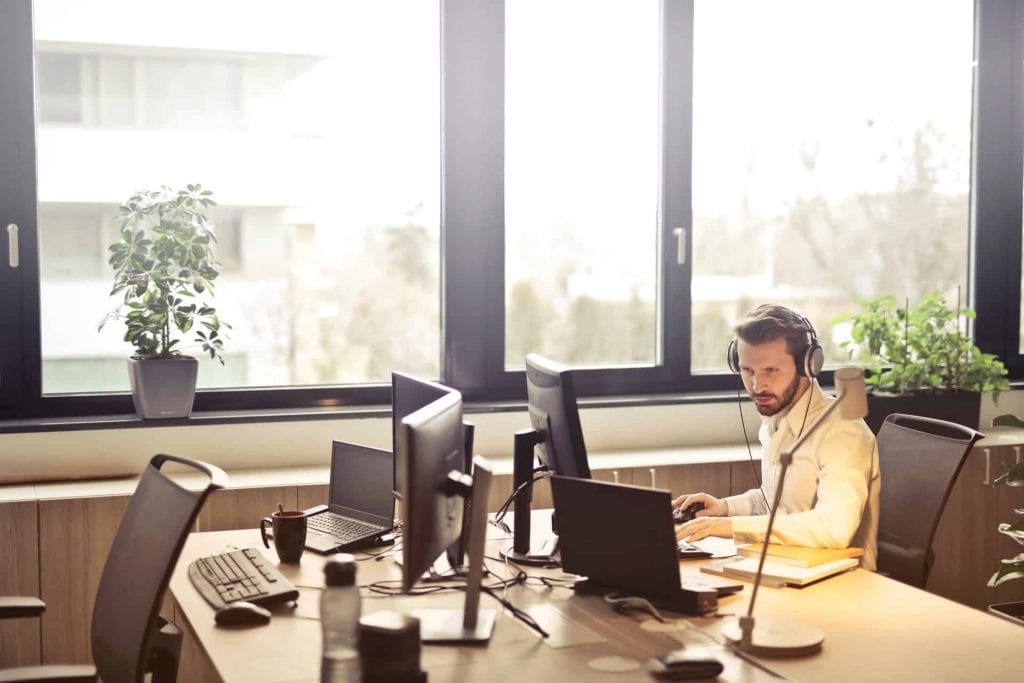 Een man in een kantooromgeving draagt een headset en werkt geconcentreerd aan meerdere computers. Op de achtergrond zijn planten zichtbaar, wat bijdraagt aan een frisse en productieve werksfeer.