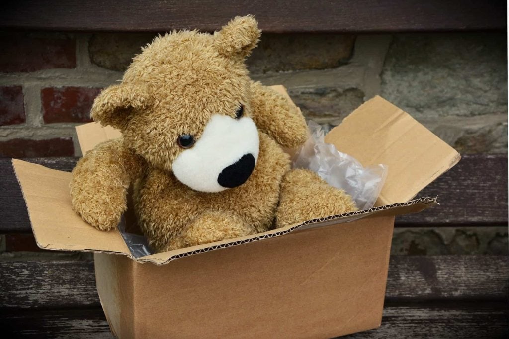 Een schattige teddybeer zit in een open kartonnen doos, omringd door plastic beschermmateriaal. De beer heeft een zachte, bruine vacht en een vriendelijke uitstraling.