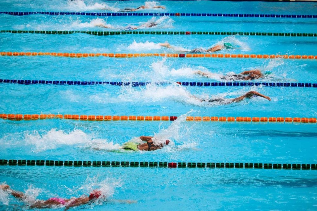 Zwemwedstrijd met meerdere zwemmers in verschillende banen in een zwembad, met spetters water en kleurrijke richtlijnen. Deze afbeelding benadrukt de dynamiek en competitie in de zwemsport.