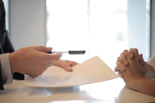 Een hand die een pen vasthoudt, terwijl een andere hand met gevouwen vingers op een tafel ligt, met een document in het zicht. Deze afbeelding illustreert een zakelijke of juridische bespreking.