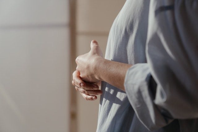 Een vrouw staat met haar handen samengevouwen, gekleed in een losse, lichtblauwe blouse. De zachte verlichting benadrukt een gevoel van rust en contemplatie.