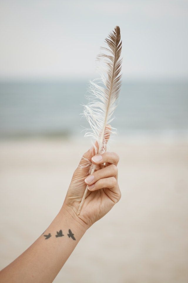 Een hand houdt een delicate veer vast met de zee op de achtergrond, terwijl de arm een tatoeage van vogels toont. Deze afbeelding straalt een gevoel van vrijheid en verbinding met de natuur uit.