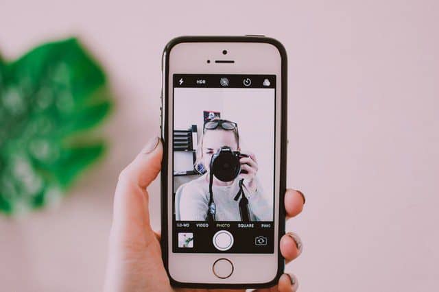 Een vrouw maakt een selfie met een smartphone terwijl ze een camera vasthoudt, met een groene plant op de achtergrond. Deze afbeelding illustreert de combinatie van moderne technologie en fotografie.