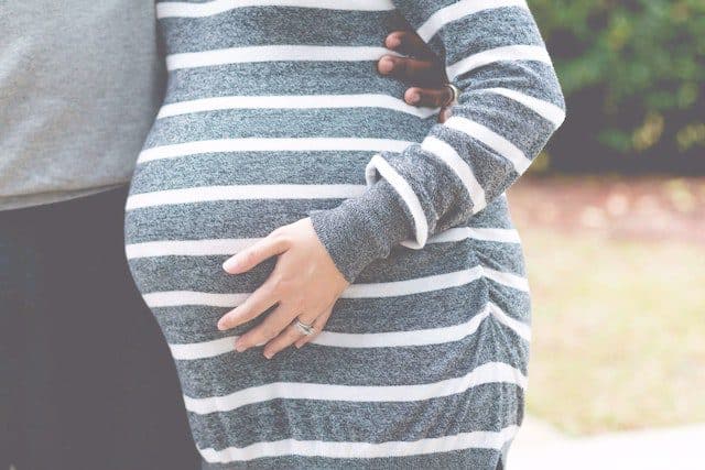 Zwangere vrouw in gestreepte jurk die haar buik vasthoudt, terwijl een hand van een partner steun biedt. De afbeelding straalt liefde en verbondenheid uit.