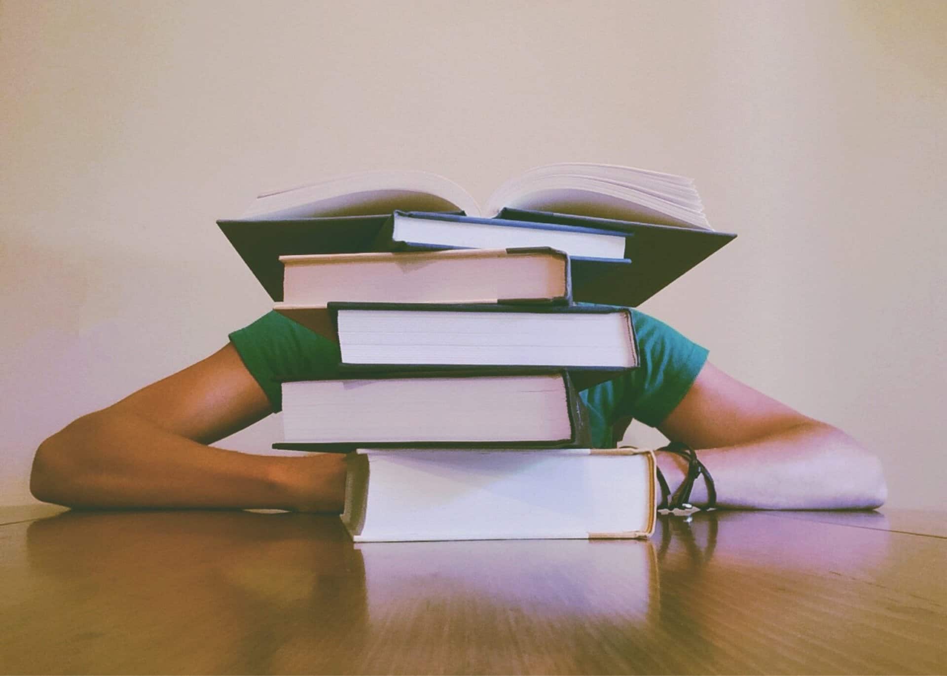 Stapels boeken met een open boek bovenop, terwijl een vrouw haar armen op de tafel steunt. De afbeelding symboliseert studeren en kennisverwerving.