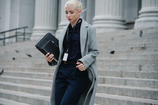 Vrouw in een stijlvolle grijze jas en donkere kleding, die een map vasthoudt terwijl ze de trappen van een gebouw afloopt. De afbeelding straalt een professionele en zelfverzekerde uitstraling uit.