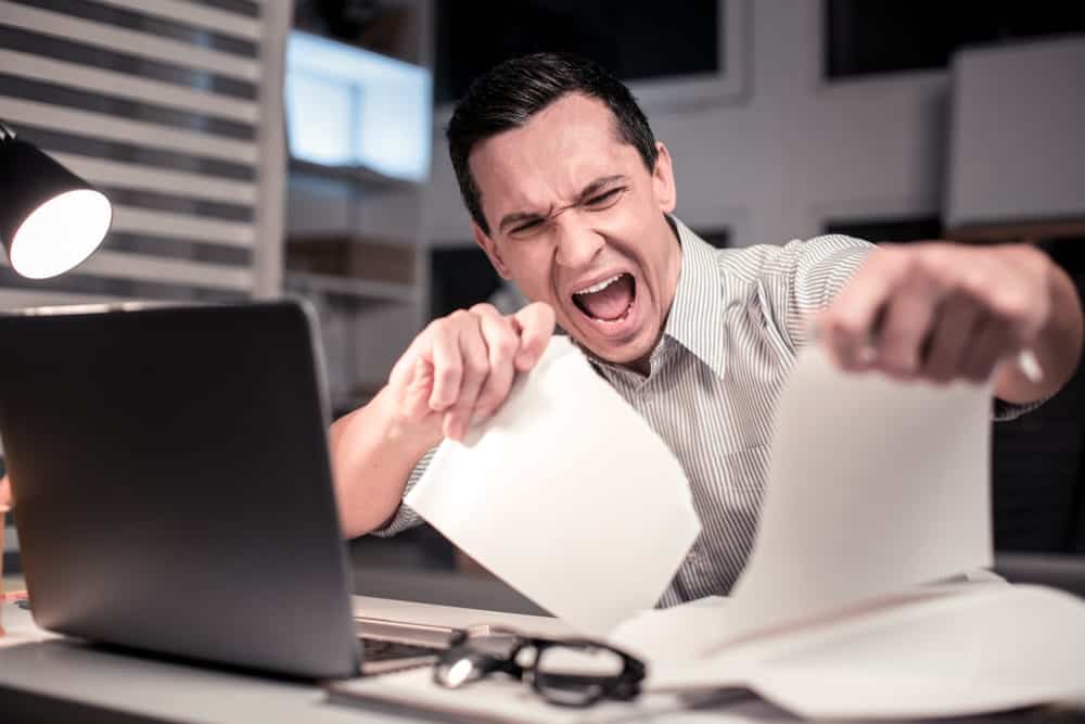 Een gefrustreerde man scheurt papieren terwijl hij voor een laptop zit, wat wijst op stress of woede over werkgerelateerde taken. De scène vindt plaats in een kantooromgeving met een bureau en een bureaulamp.