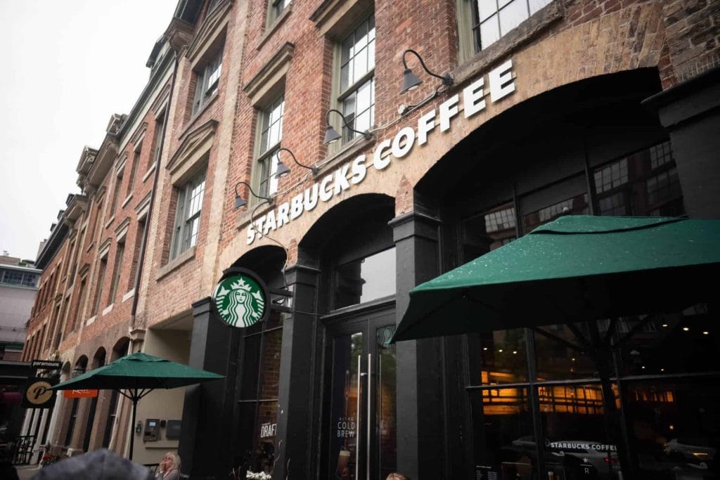 Starbucks-vestiging met een kenmerkende groene logo en terras met parasols. De gevel is gebouwd van bakstenen en heeft grote ramen, wat een uitnodigende sfeer creëert voor koffieliefhebbers.